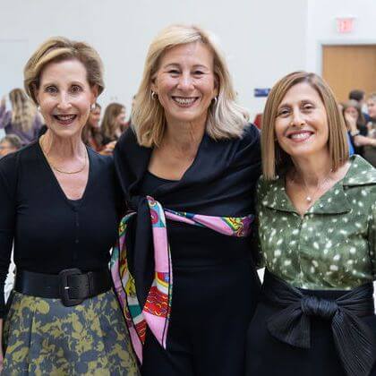 Three women smiling and standing together at an indoor event. They are dressed in stylish outfits, and there are people seated in the background, suggesting a social gathering or function.