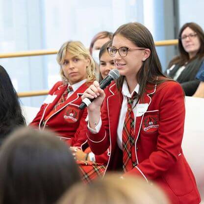 A student in a red blazer and plaid tie speaks into a microphone at an indoor event, while other similarly dressed students and attendees listen in the background.