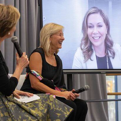 Two women sit and talk into microphones during an event, while a third woman smiles on a large video screen behind them, participating virtually. All three appear engaged and happy.