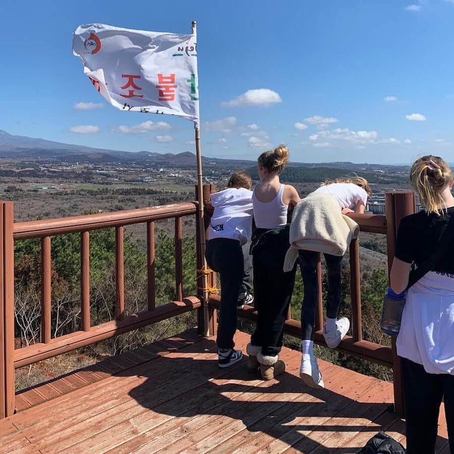 Four children stand and lean over a wooden lookout platform with a flag featuring Korean writing. The view shows a wide landscape with trees, fields, and a mountain under a blue sky with scattered clouds.