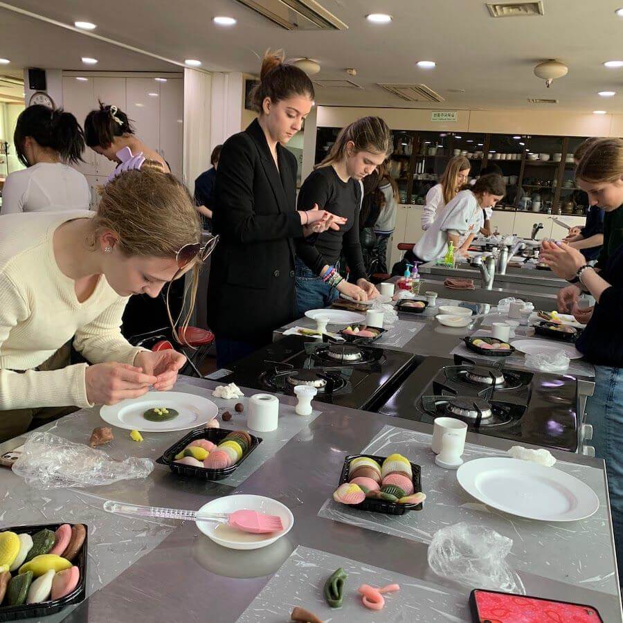 Several people concentrate on making colorful confections at a workshop, working at tables with trays of sweets and kitchen tools. The room is bright and filled with natural light.