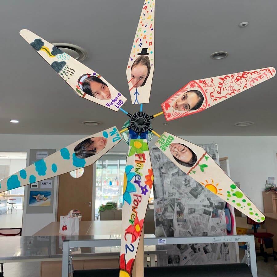 A colorful handmade windmill with seven blades, each decorated with bright patterns and children's photos. The windmill stands indoors in a classroom with art supplies, tables, and artwork in the background.