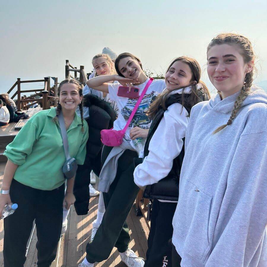 Five young women pose and smile together outdoors on a sunny day. Four wear casual sweatshirts and one wears a bright green shirt. They stand on a wooden deck, with some holding water bottles.