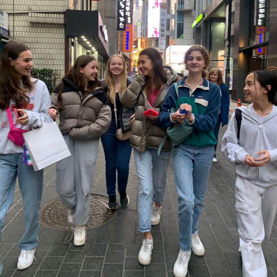 Six teenage girls walk together down a city street, smiling and chatting. They wear casual clothing and jackets. Neon signs and tall buildings line the background, giving an urban vibe.