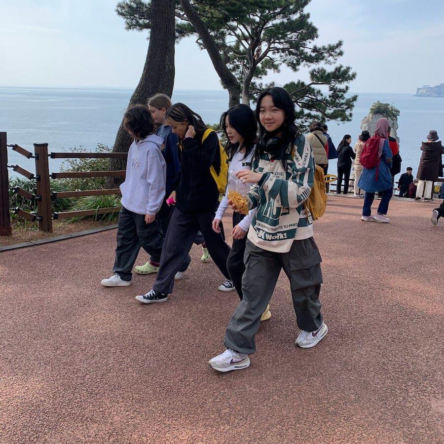 A group of young people walk along a paved path beside the sea, surrounded by trees and other visitors. Some wear hoodies and backpacks, enjoying a scenic outdoor area with water and cliffs visible in the background.