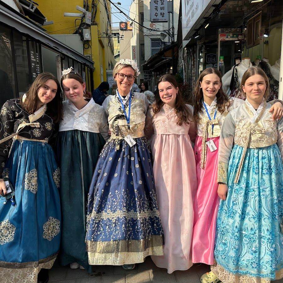 Six people stand closely together smiling, wearing colorful traditional Korean hanbok dresses, on a sunny street lined with shops and signs in Korean.