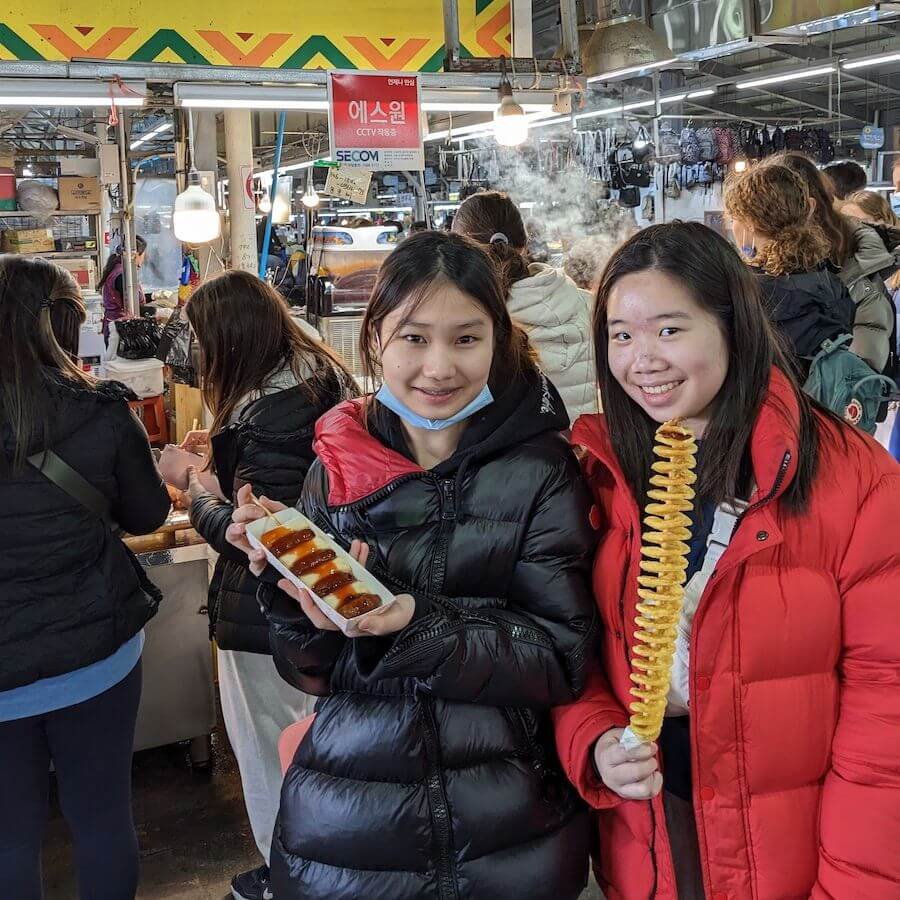 Two young women in winter jackets smiling at a busy indoor market. One holds Korean rice cakes on a stick, and the other holds a spiral-cut fried potato skewer. People and food stalls are visible in the background.