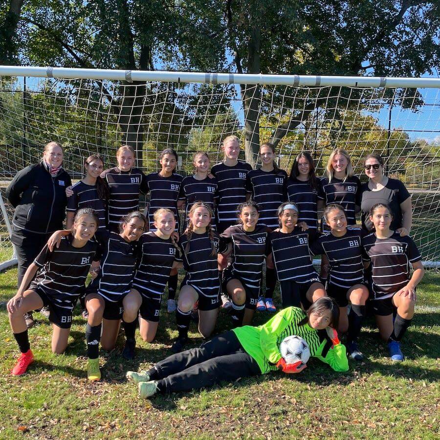 A girls’ soccer team in black uniforms poses in front of a goal with two coaches. One player in a green goalkeeper uniform lies on the grass in front, holding a soccer ball and smiling. Trees and blue sky are visible behind them.