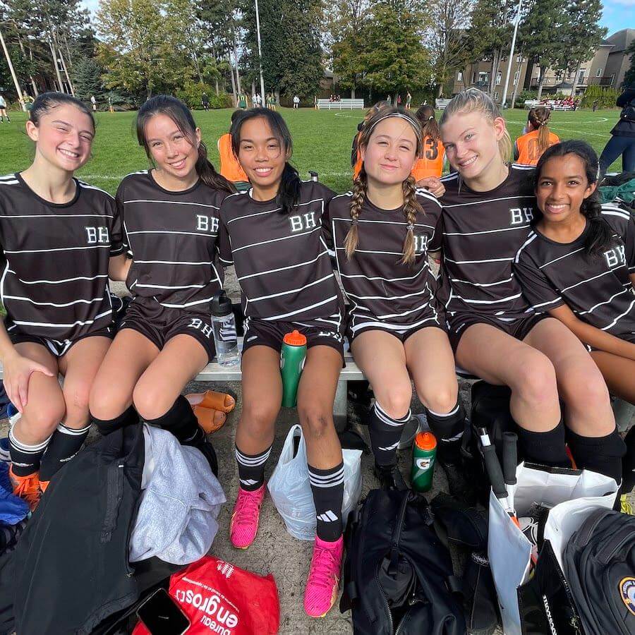 Six teenage girls in matching black striped soccer uniforms sit on a bench outdoors, smiling at the camera. They have water bottles and sports bags at their feet, and trees and other players are visible in the background.