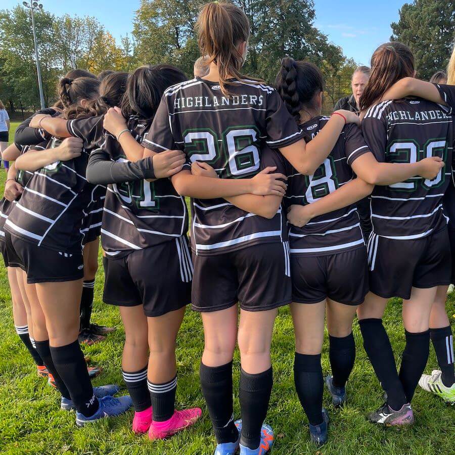 A girls' soccer team in black "Highlanders" uniforms stands in a huddle on a grassy field, arms around each other's shoulders, preparing for their game.