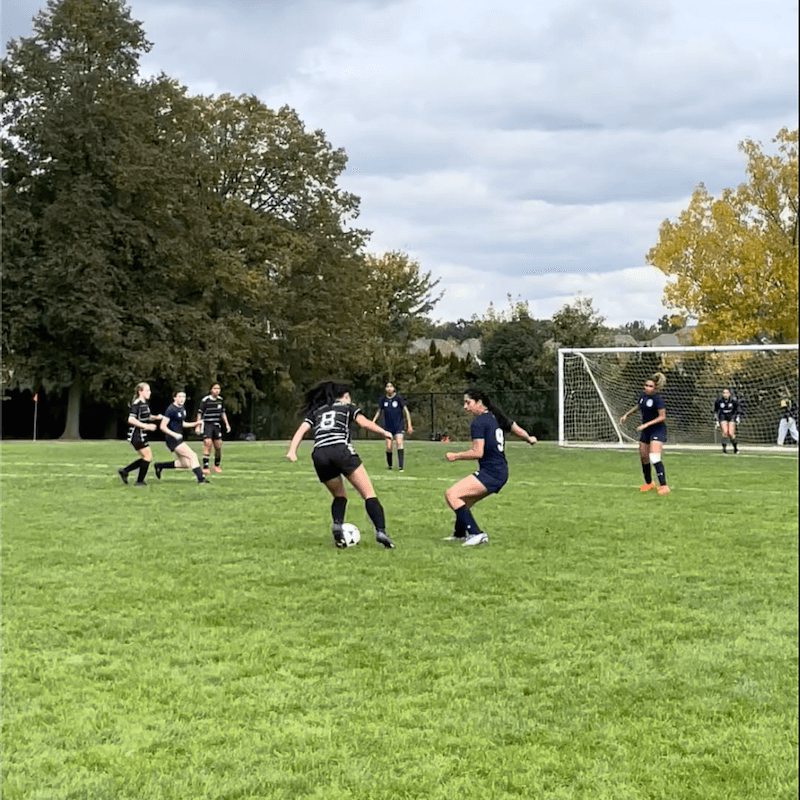 A girls’ soccer match in progress on a grassy field; one player in a black uniform dribbles the ball while a defender in a navy uniform blocks. Other players and a goalpost are visible in the background under cloudy skies.