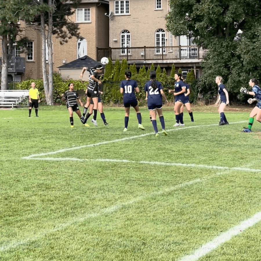 A soccer player in a striped jersey heads the ball in front of the goal while surrounded by opponents in navy blue uniforms, as the goalkeeper and other players watch intently. A referee stands in the background.