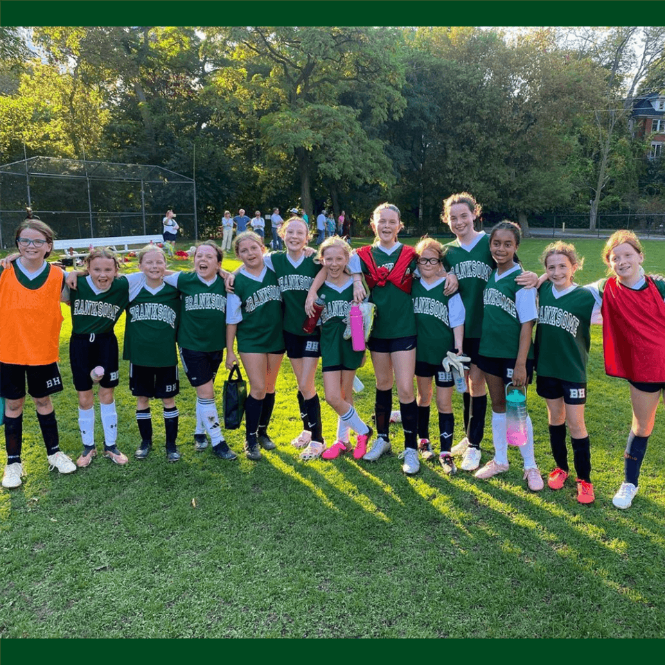 A girls’ soccer team stands side by side on a grassy field, smiling at the camera. They wear green Saintrose jerseys and soccer gear, with trees and a baseball backstop visible in the background.
