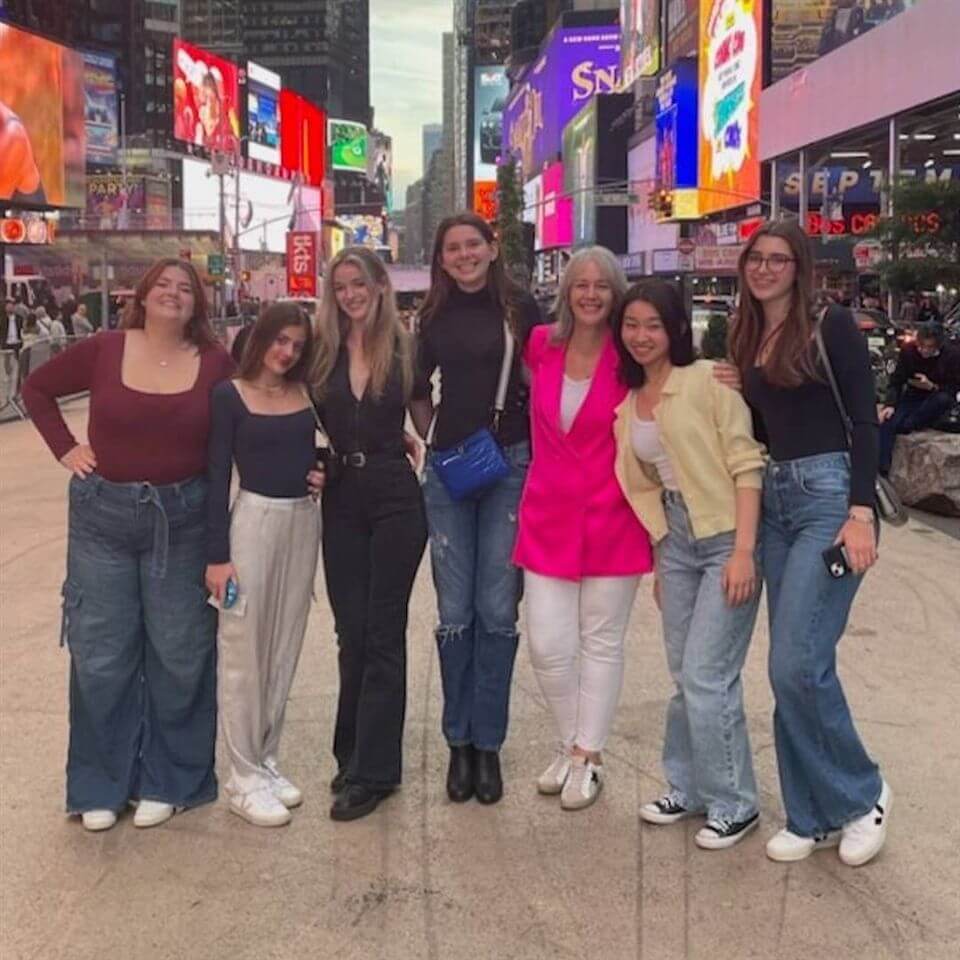 Seven women stand together and smile for a group photo in a busy, brightly lit urban area with colorful billboards, likely Times Square. They wear casual outfits, with one in a bright pink blazer and white pants.