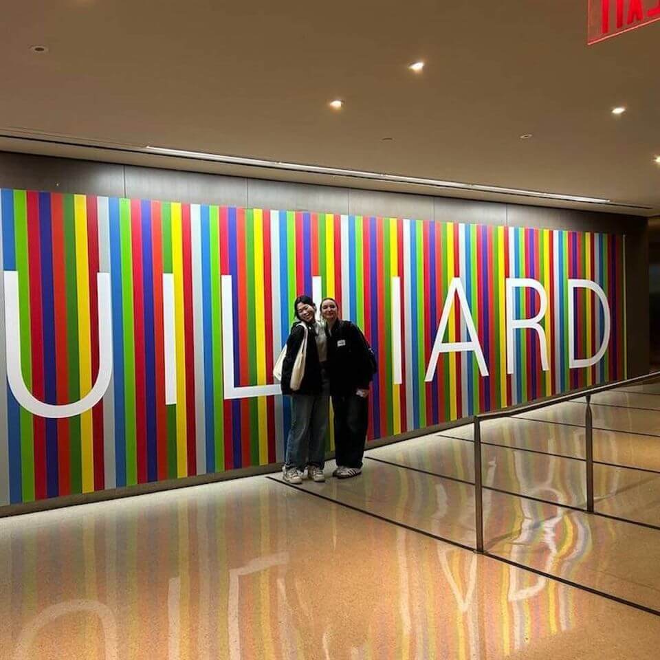 Two people pose and smile in front of a colorful striped wall with large white letters spelling “Juilliard.” The hallway is brightly lit with a shiny floor and a railing along the side.