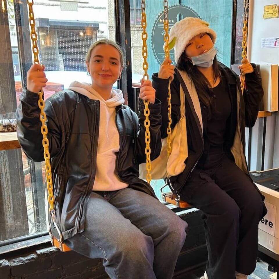 Two people sit on indoor swings by a large window. One person wears a leather jacket and hoodie; the other wears a bucket hat, face mask under chin, and a two-toned jacket. Both appear relaxed and are looking at the camera.