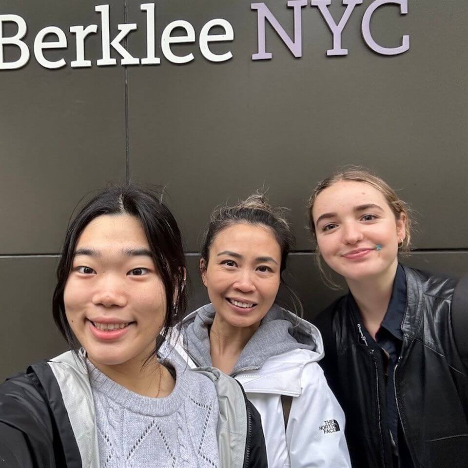 Three people smile for a selfie in front of a wall with the words "Berklee NYC" on it. They are dressed in casual clothing and appear happy to be together.
