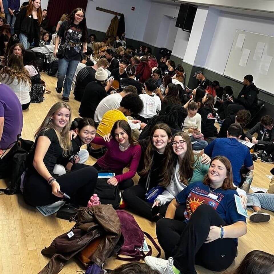 A large group of students sit on a gymnasium floor in clusters, talking and smiling. In the foreground, six young women pose together, smiling at the camera. The room is lively and filled with people.