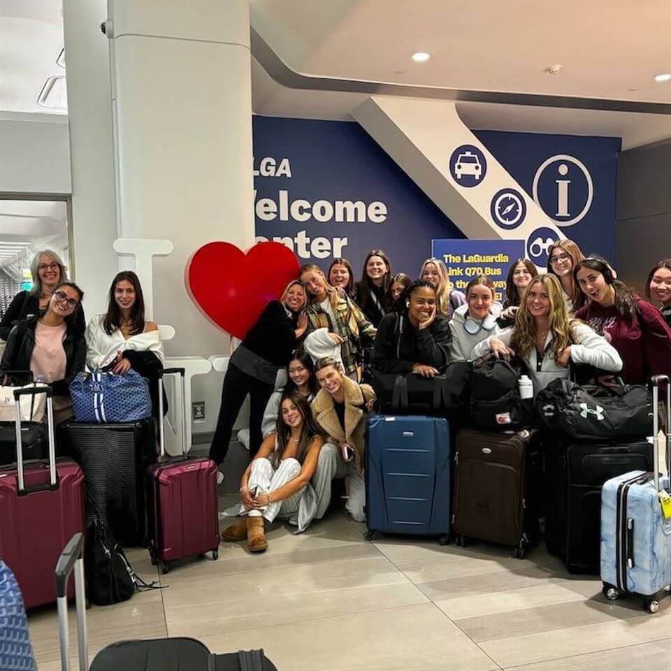 A group of people with suitcases pose and smile in front of an “I ❤️” sign at an airport welcome center, with travel signs and luggage carts visible in the background.