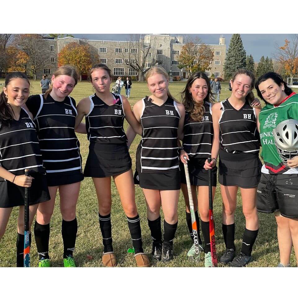 A group of seven field hockey players in black uniforms stands together on a grassy field, smiling at the camera. A school building and autumn trees appear in the background.