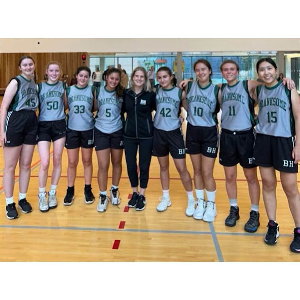 Nine young women in basketball uniforms, standing arm-in-arm on a gym court, smiling at the camera. Most are wearing gray jerseys with "Branksome" and various numbers. A coach stands in the center.