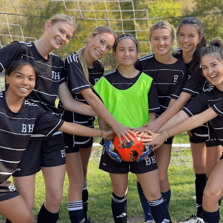 Six smiling teenage girls in black soccer uniforms stand in front of a goal, with one player wearing a neon green vest. They all have their hands on an orange soccer ball, showing team unity.