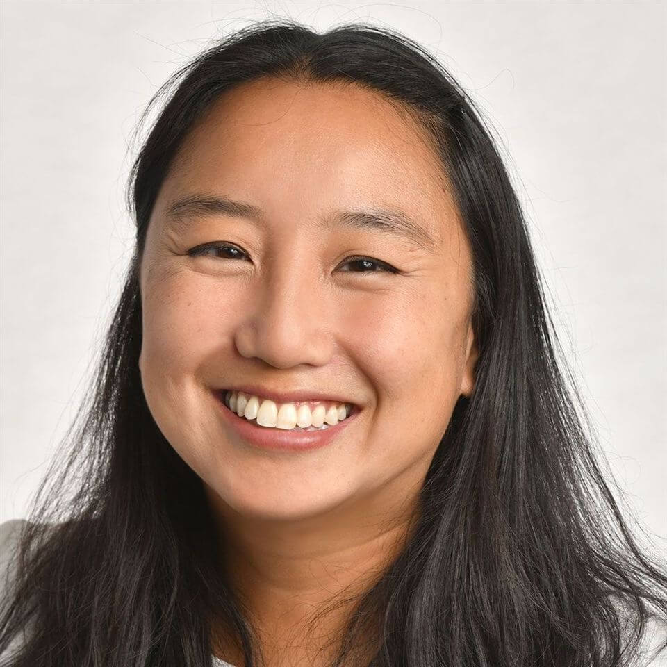 A woman with long black hair smiles at the camera. She is against a plain, light-colored background and is wearing a white top.
