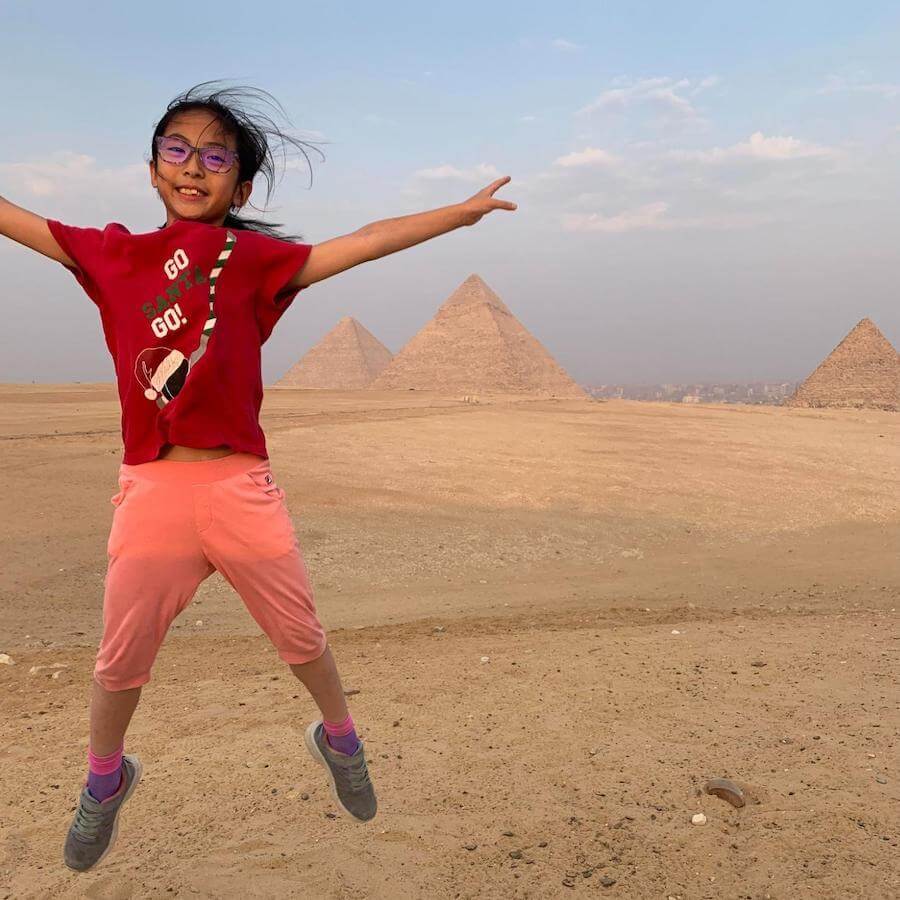 A smiling girl in a red t-shirt and pink shorts jumps with arms outstretched in front of the Pyramids of Giza, Egypt, on a sunny day with a partly cloudy sky.