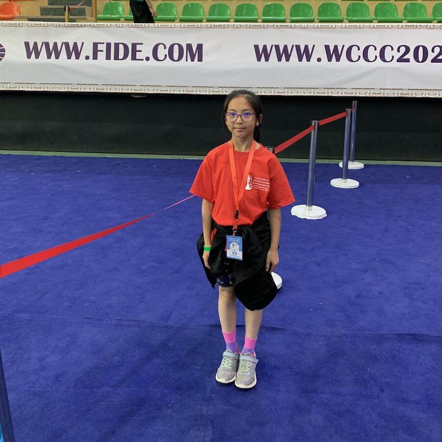 A young girl wearing glasses, a red shirt, shorts, and sneakers stands on a blue carpet in front of a barrier. Behind her is a white banner with "FIDE.COM" and "WCCC202" visible.