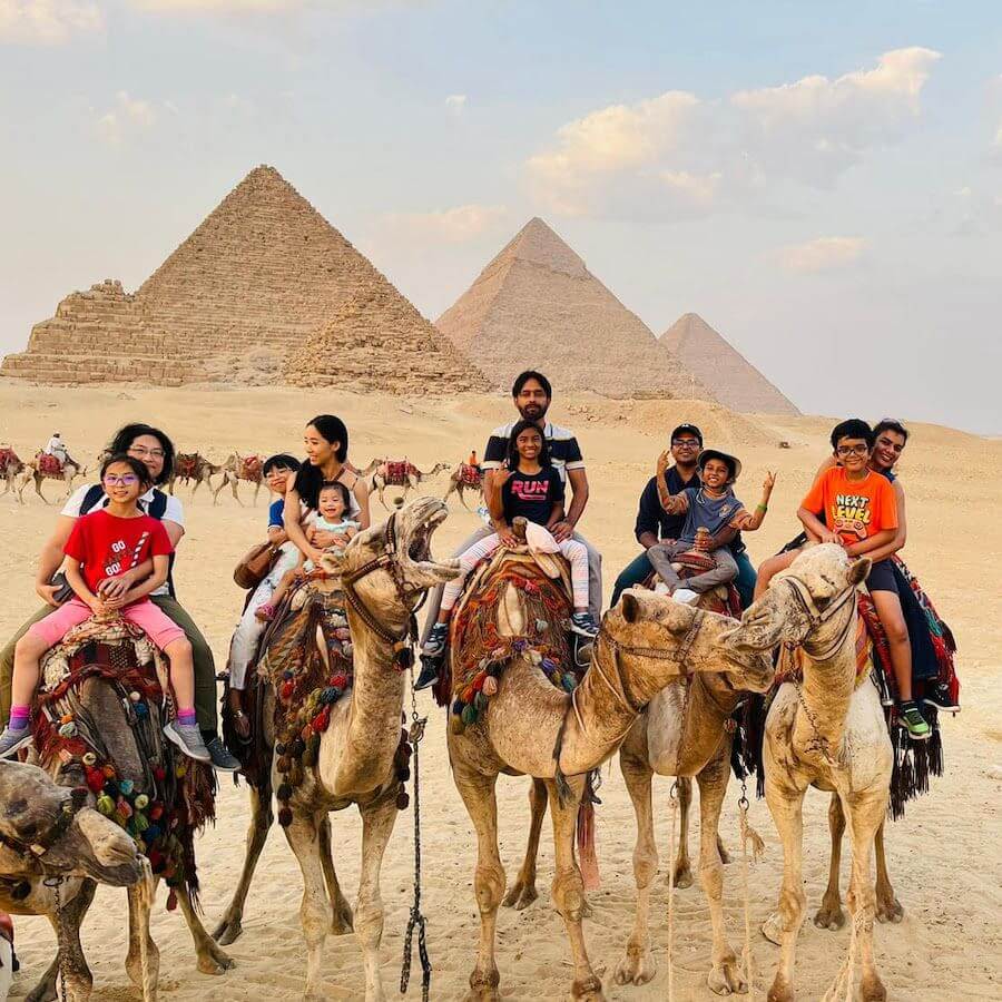 A group of people, including children and adults, are riding camels in front of the Pyramids of Giza in Egypt on a sunny day. The desert landscape and pyramids are visible in the background.