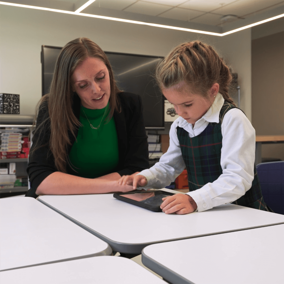 A teacher sits beside a young girl in a plaid school uniform, guiding her as she uses a tablet at a classroom table. The classroom has books and modern décor in the background.