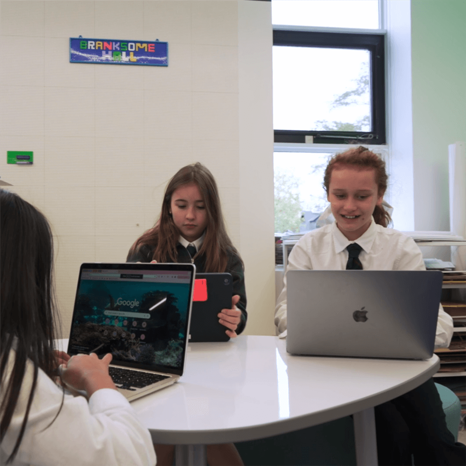Three students in school uniforms sit around a white table using laptops and tablets in a classroom with large windows and a Branksome Hall sign on the wall.