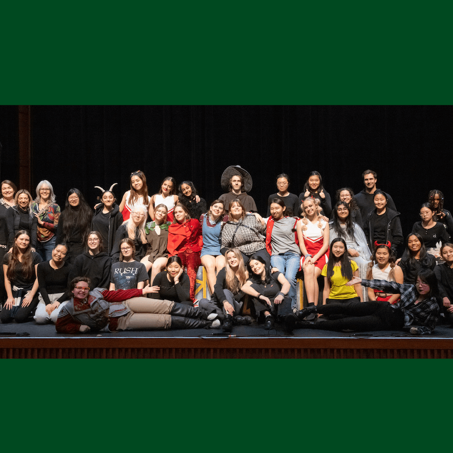 A large group of people, many in costumes, sit and stand together on a stage, smiling at the camera. The background is dark, highlighting the group in the foreground.