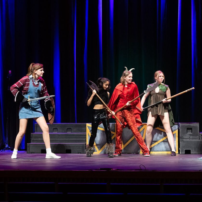 Four people in costume stand on stage, each holding prop weapons and looking alert. The background features vertical lights in blue and purple, and stage platforms behind them. The scene suggests a theatrical performance or play.