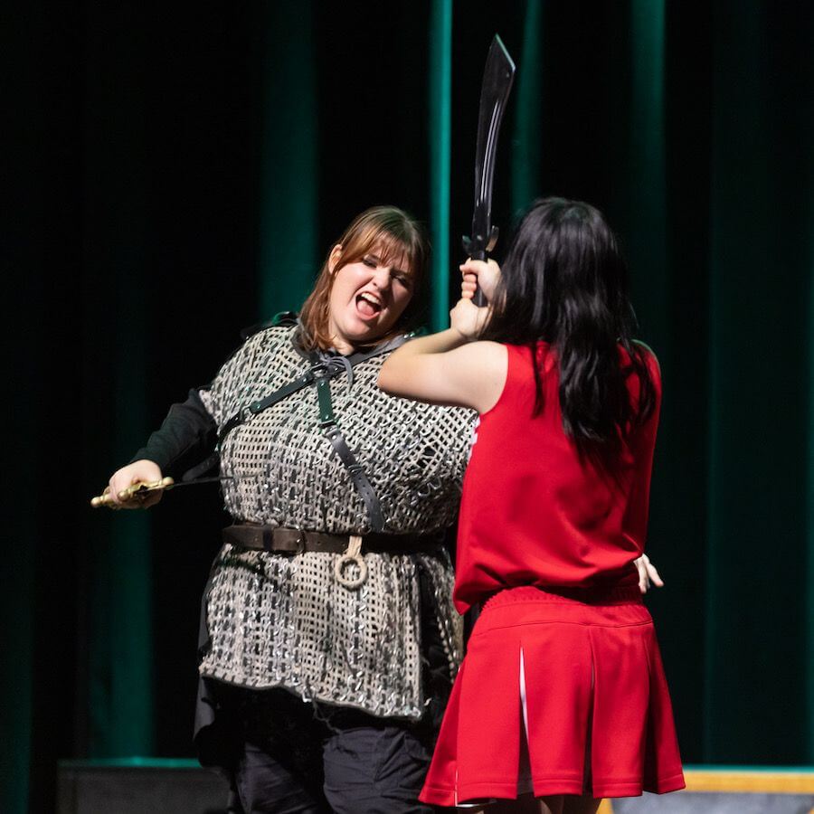 Two people are performing on stage; one in a red dress raises a prop sword while the other, dressed in medieval-style attire, reacts dramatically with their tongue out. Green curtains are in the background.