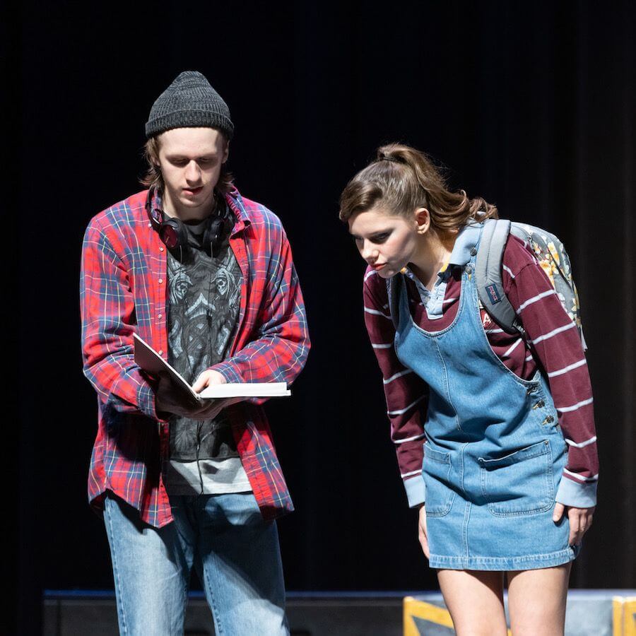 Two young people perform on stage. One wears a beanie and plaid shirt, holding an open book. The other, in a denim dress and striped shirt with a backpack, bends forward to look at the book. Both appear focused.