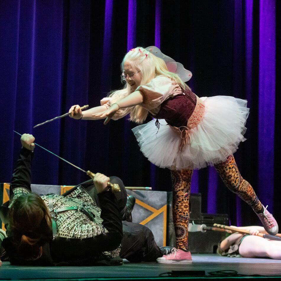 A person in a fairy costume, with wings and a tutu, playfully points a wand at someone lying on the stage holding a sword, set against a background of dark blue curtains.