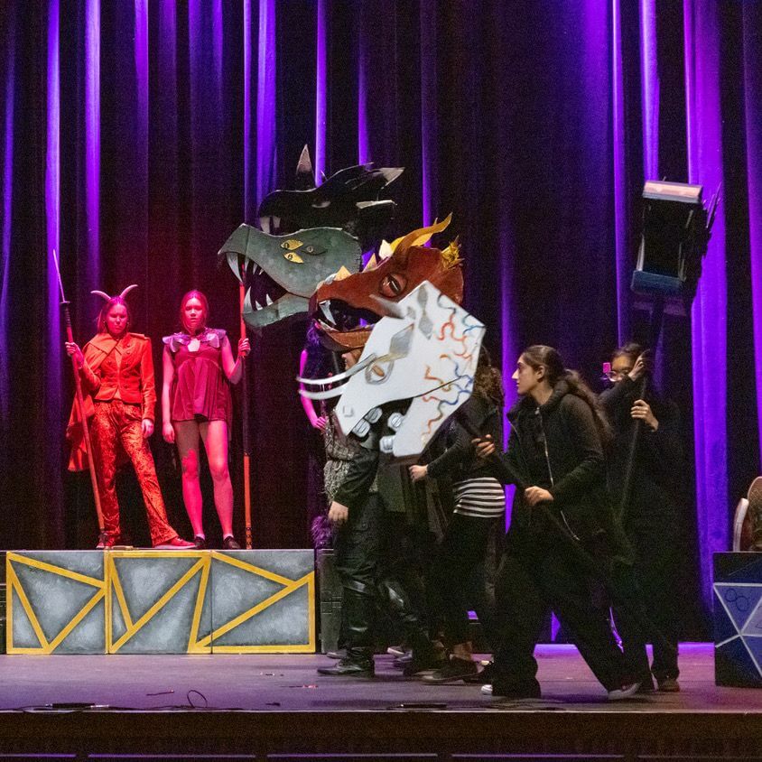 A group of performers on stage, some dressed in red costumes with spears, others operating large dragon head puppets. Purple stage lighting and geometric set pieces decorate the scene.