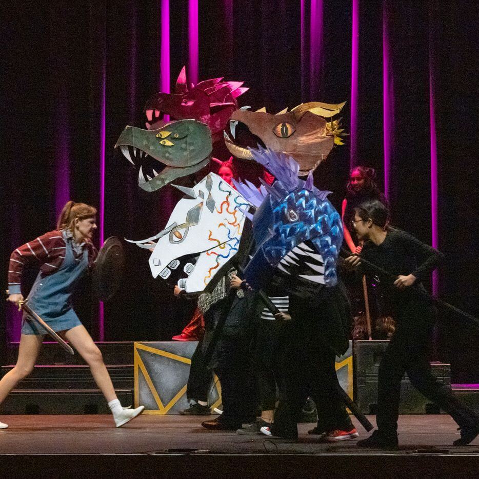Five people on stage perform with colorful dragon head puppets. One person holds a shield, facing the puppet heads, as others operate the dragons. The background features vertical pink-purple stage lights.