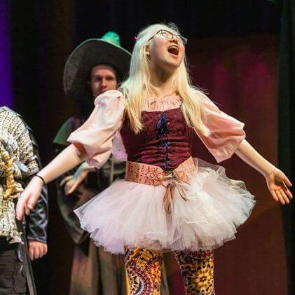 A young person in a stage costume, including a tutu and patterned leggings, stands with arms wide open and mouth agape, appearing to sing or perform. Two other people in costumes are visible in the background.