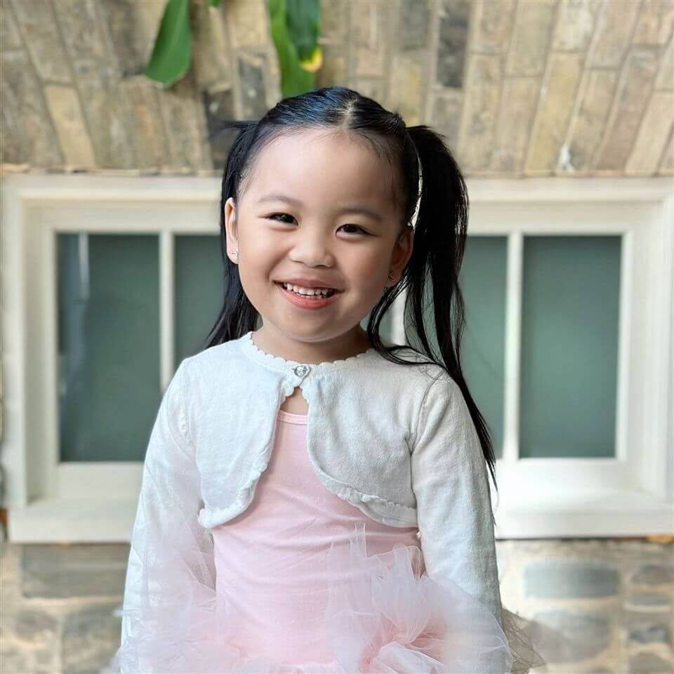 A young girl with long black hair in pigtails smiles at the camera. She is wearing a light pink dress with a white cardigan. Behind her is a window set in a stone wall.