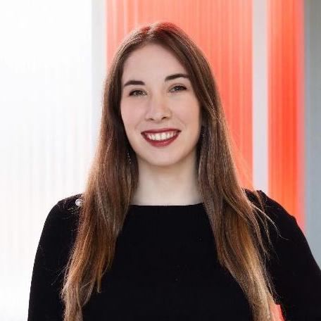 A woman with long straight brown hair and fair skin smiles at the camera. She is wearing a black top and standing in front of a background with vertical orange and white panels.
