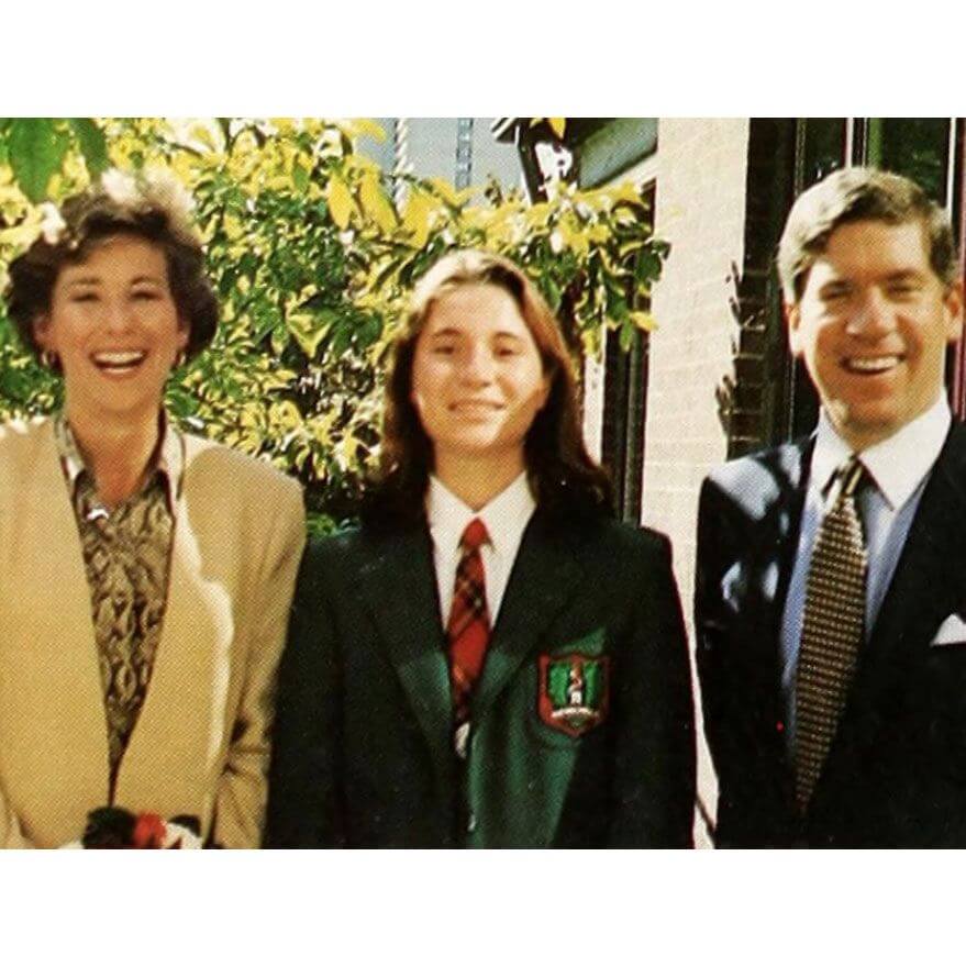 Three people, two adults in business attire and a young person in a green school blazer and red striped tie, stand smiling outdoors in sunlight with greenery and a building in the background.