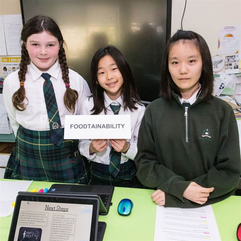 Three schoolgirls stand at a presentation table. One holds a sign that says "FOODSTAINABILITY." They wear uniforms, and a laptop, mouse, and documents are on the table in front of them.