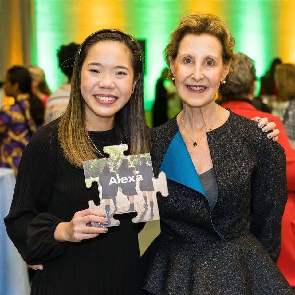 Two women smiling at a social event; the woman on the left holds a large puzzle piece with the name "Alexa" and a photo of people walking. The background features people mingling and green-yellow lighting.