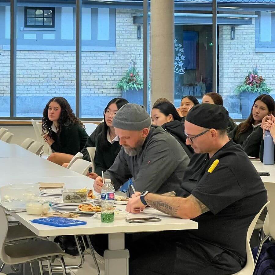 Two men sit at a table, taking notes and tasting food, while a group of people, mostly women, sit attentively in the background. The setting appears to be a modern classroom or meeting space with large windows.