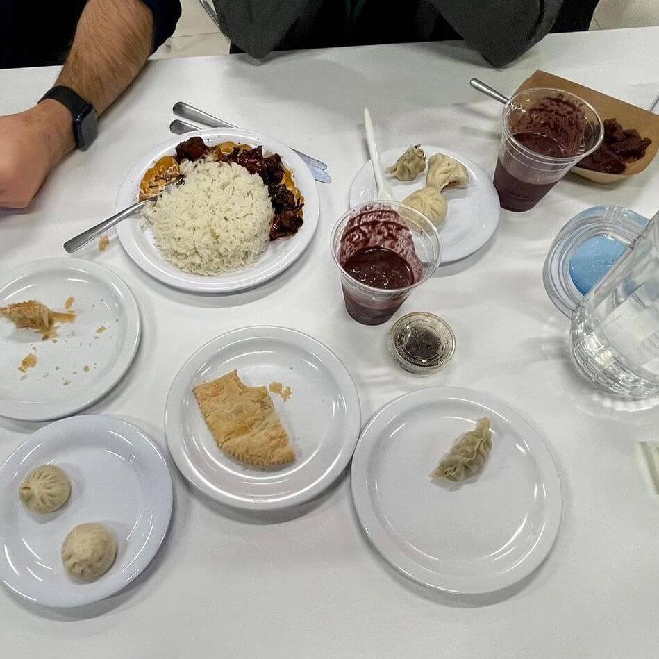 A table with several plates of food, including rice with meat, dumplings, a pastry, cups with chocolate dessert, a small container with sauce, and a jug of water. Three people's arms are visible at the table.