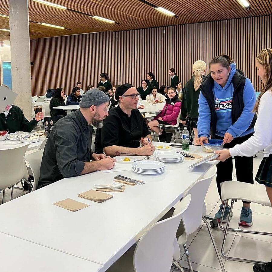A girl in a blue hoodie serves food to two men seated at a white table in a busy cafeteria, while groups of people talk and eat in the background.