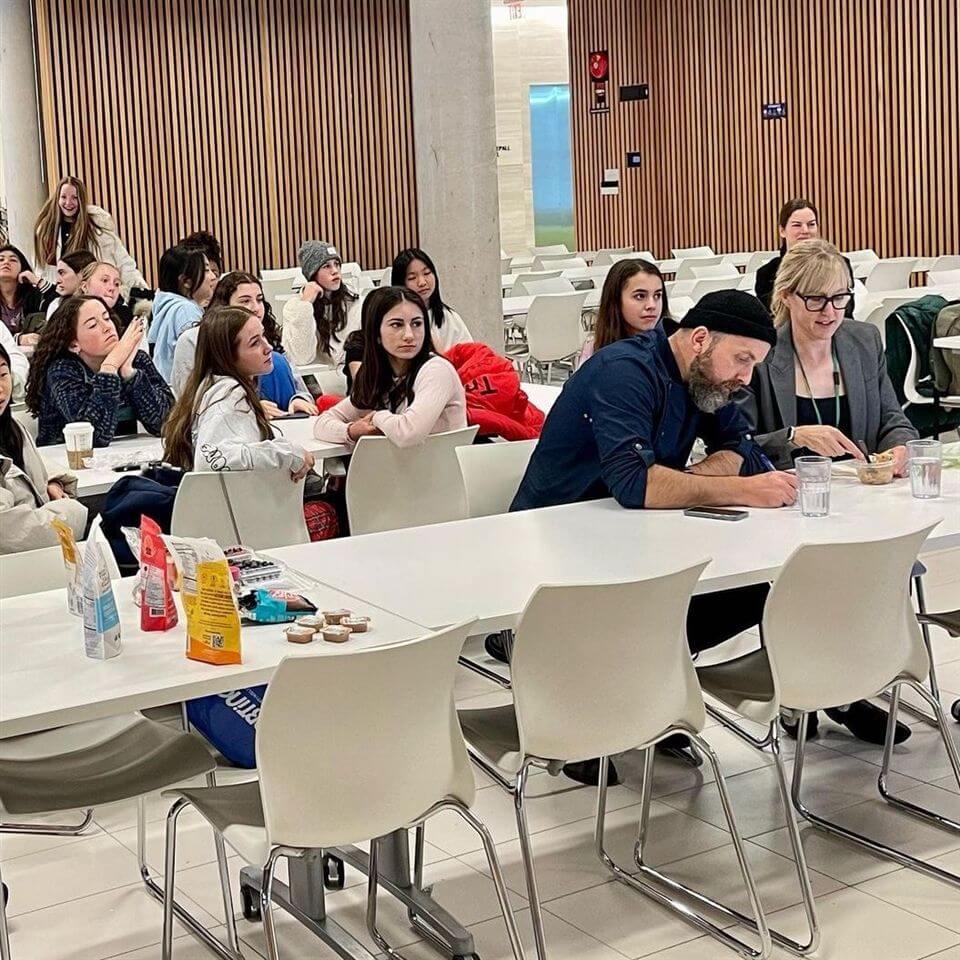 A group of people sit at white tables in a modern, indoor space. Some are listening attentively, while two in the front write notes. There are snacks and drinks on the tables, and a wood-paneled wall is in the background.