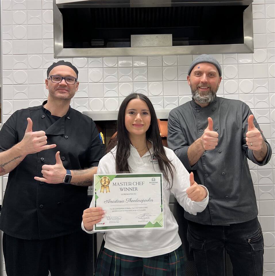 Three people stand in a kitchen. The woman in the middle holds a "Master Chef Winner" certificate and smiles, while the two men on either side, wearing chef uniforms, give enthusiastic thumbs up.